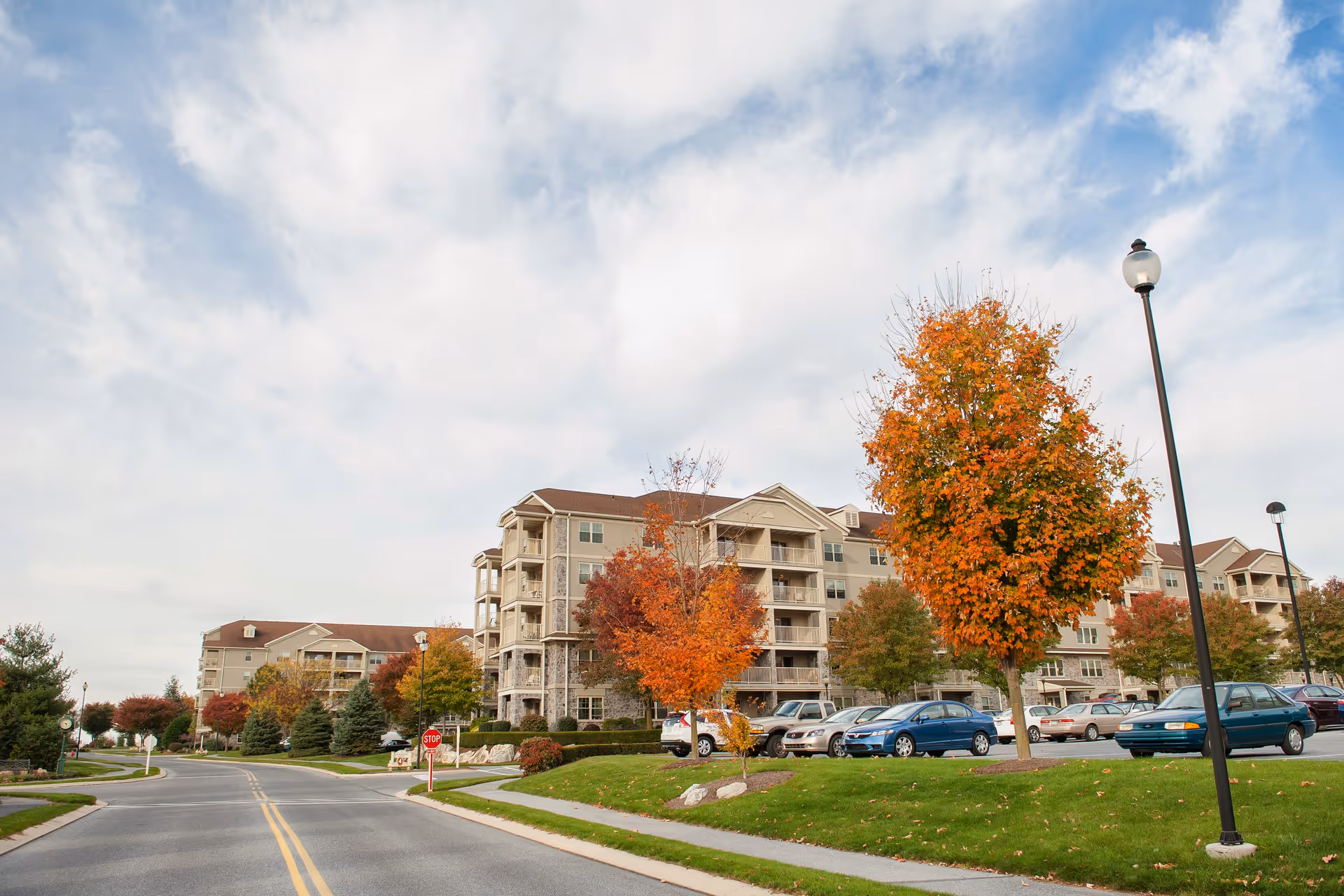 Exterior view of a multi-story senior living building with parked cars and autumn trees under a partly cloudy sky.
