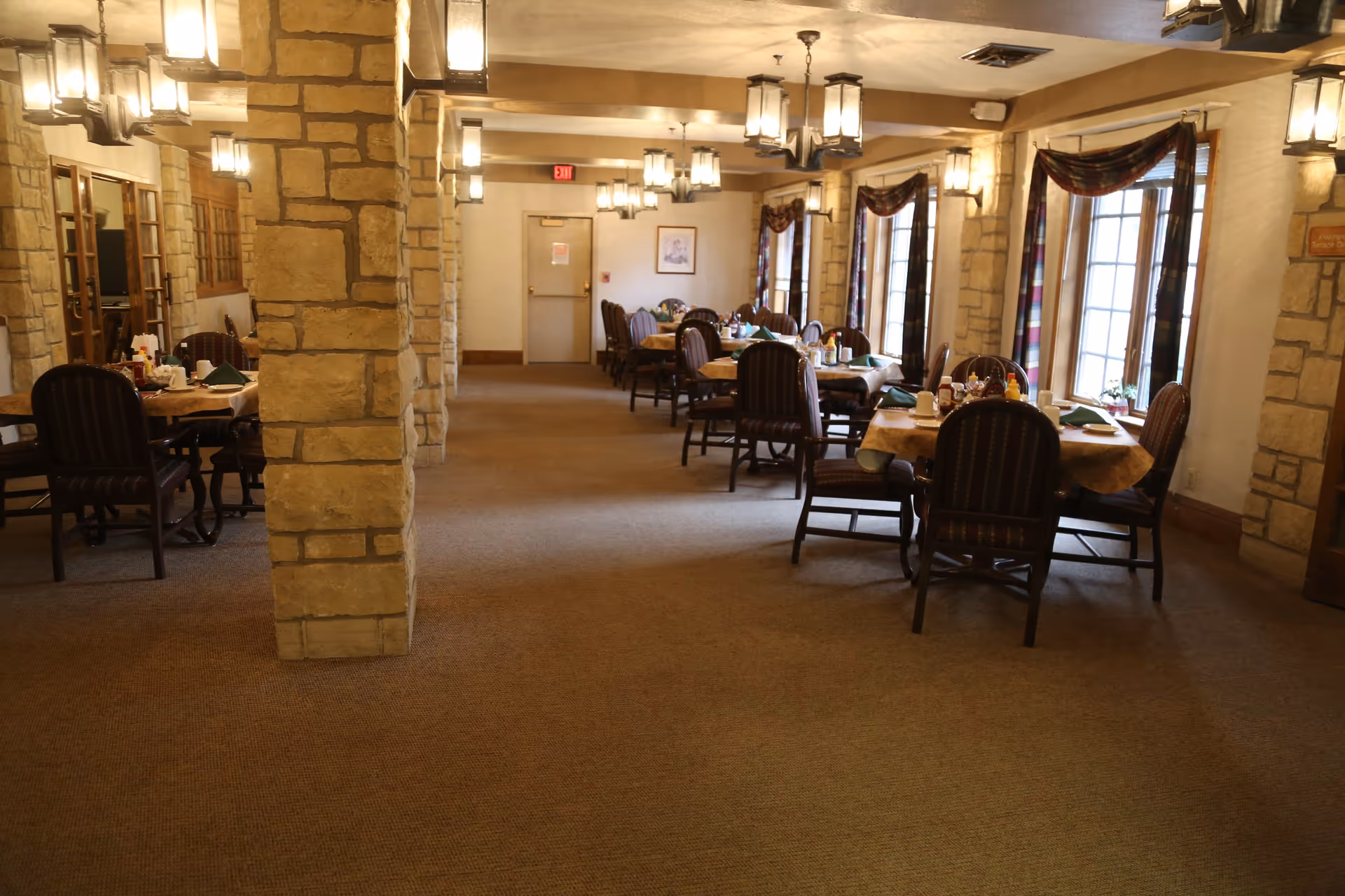 Interior view of a dining room in Cumbernauld Village featuring multiple tables with chairs arranged around them. The tables are set with tablecloths, napkins, cups, and condiments. The room has stone pillars, carpeted floor, large windows with plaid curtains, and multiple hanging light fixtures providing warm lighting.