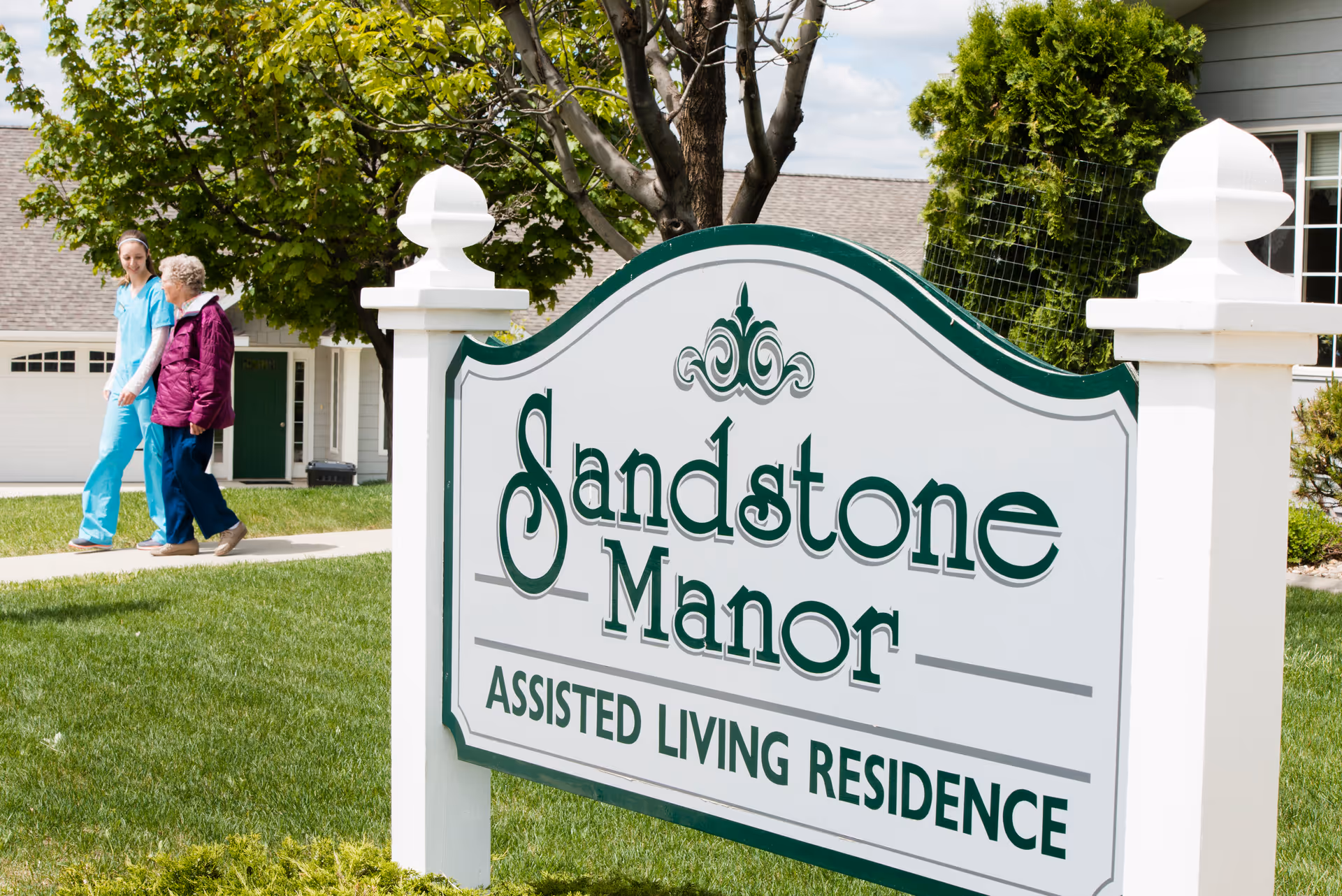 A large white and green sign reading 'Sandstone Manor Assisted Living Residence' is displayed outdoors on a grassy lawn with trees and a building in the background. To the left, a caregiver in blue scrubs walks alongside an elderly woman wearing a purple jacket on a sidewalk.