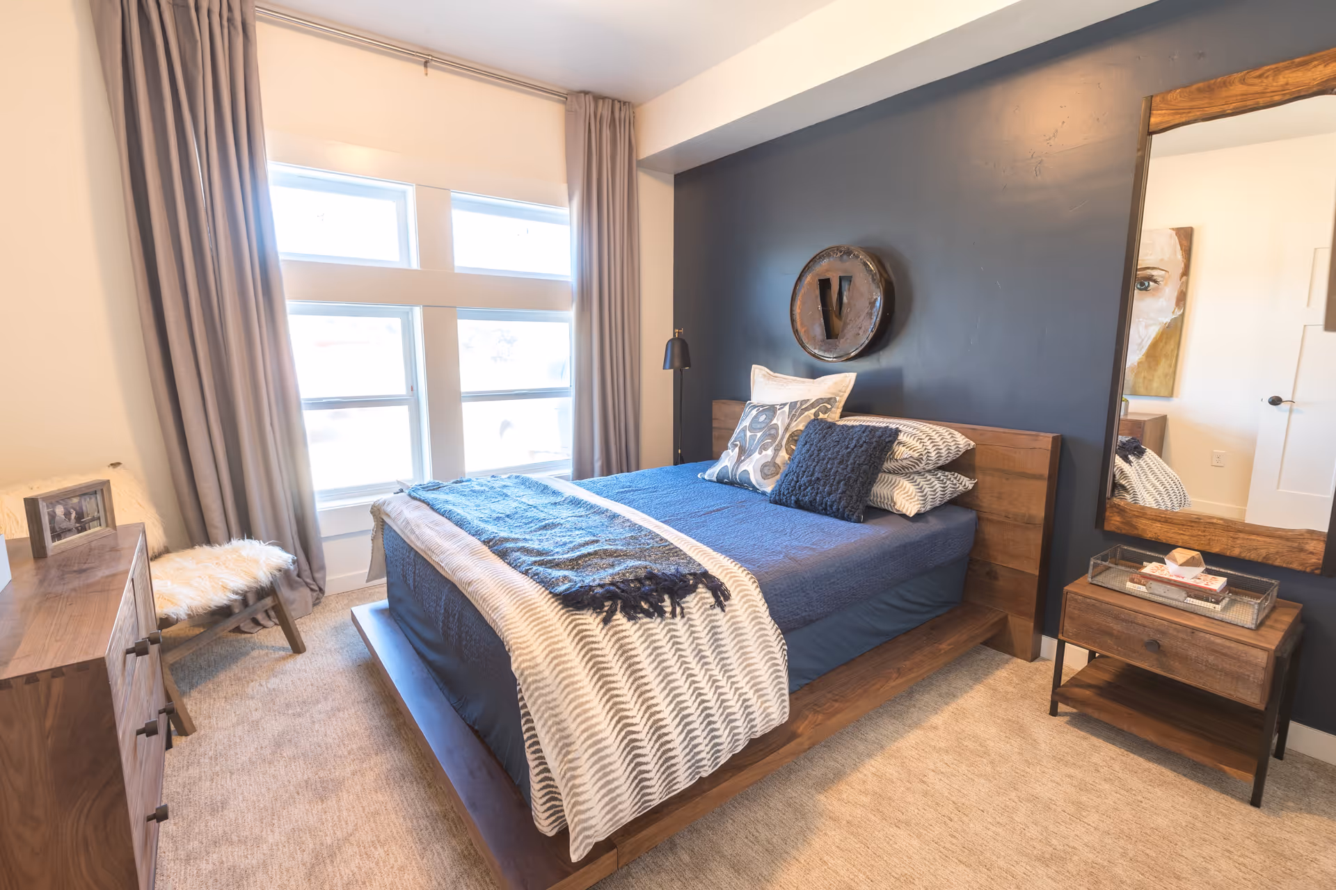A cozy bedroom with a wooden bed frame and blue bedding, including multiple pillows and a patterned throw blanket. The room features a dark accent wall behind the bed with a circular decorative piece mounted on it. There is a wooden nightstand with a drawer and a mirror above it on the right side of the bed. On the left side, there is a wooden dresser and a small chair with a white fur cushion. Large windows with gray curtains allow natural light to fill the room.