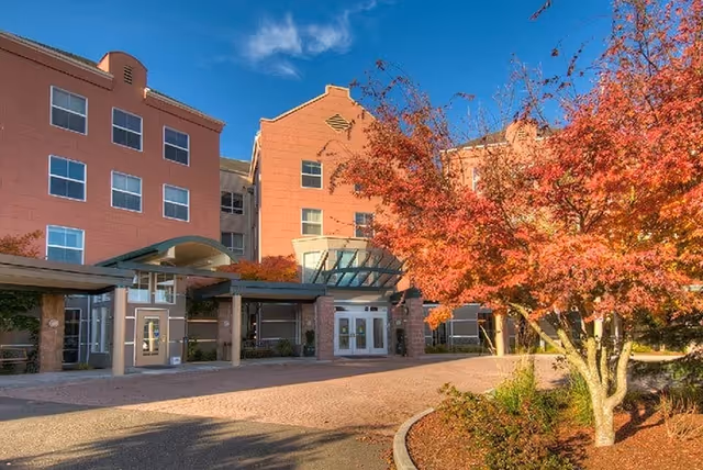 Exterior view of a multi-story retirement facility building with a covered entrance, surrounded by landscaping including a tree with red autumn leaves under a clear blue sky.