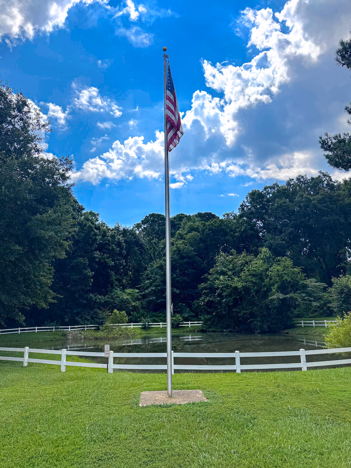 An American flag on a tall flagpole stands before a small pond and white fence with trees under a blue sky with clouds.