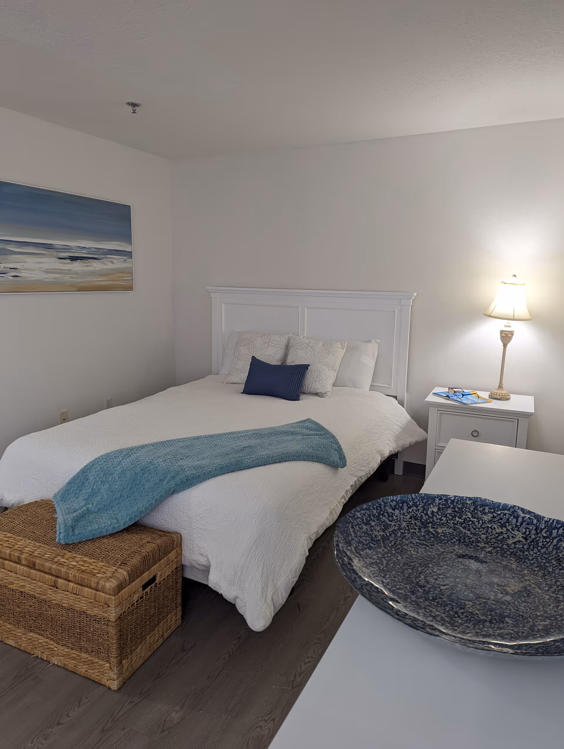 A bedroom with a white bed featuring a white headboard, white bedding, and decorative pillows including a blue one. A light blue throw blanket is draped across the foot of the bed. There is a wicker storage chest at the foot of the bed. A white nightstand with a lamp, glasses, and a book is beside the bed. A large blue decorative bowl is on a white dresser in the foreground. A painting of a beach scene hangs on the wall.