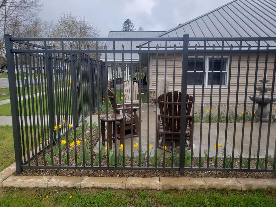 Black metal-fenced patio with Adirondack chairs, a small table, and yellow daffodils in front of a beige single-story building.