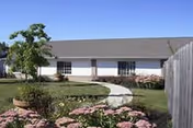 Single-story white building with a curved walkway, landscaped lawn and flower beds under a clear blue sky.