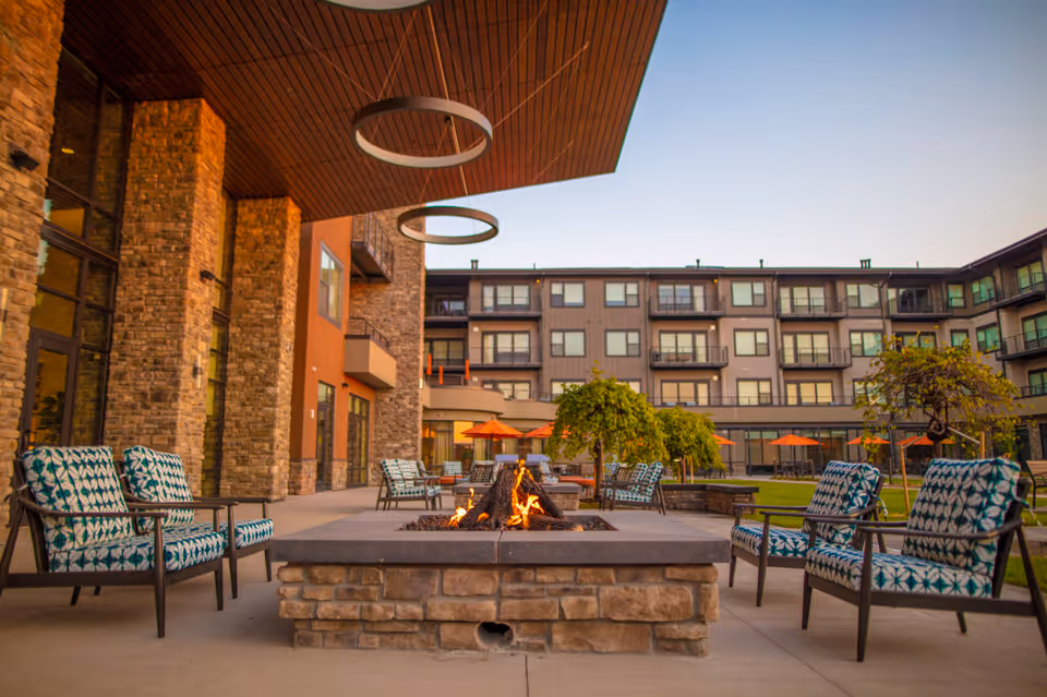Outdoor patio area at The Ridge Foothill featuring a stone fire pit surrounded by cushioned chairs with patterned upholstery. The patio is adjacent to a multi-story building with balconies and large windows. There are orange umbrellas and small trees in the background under a clear sky.