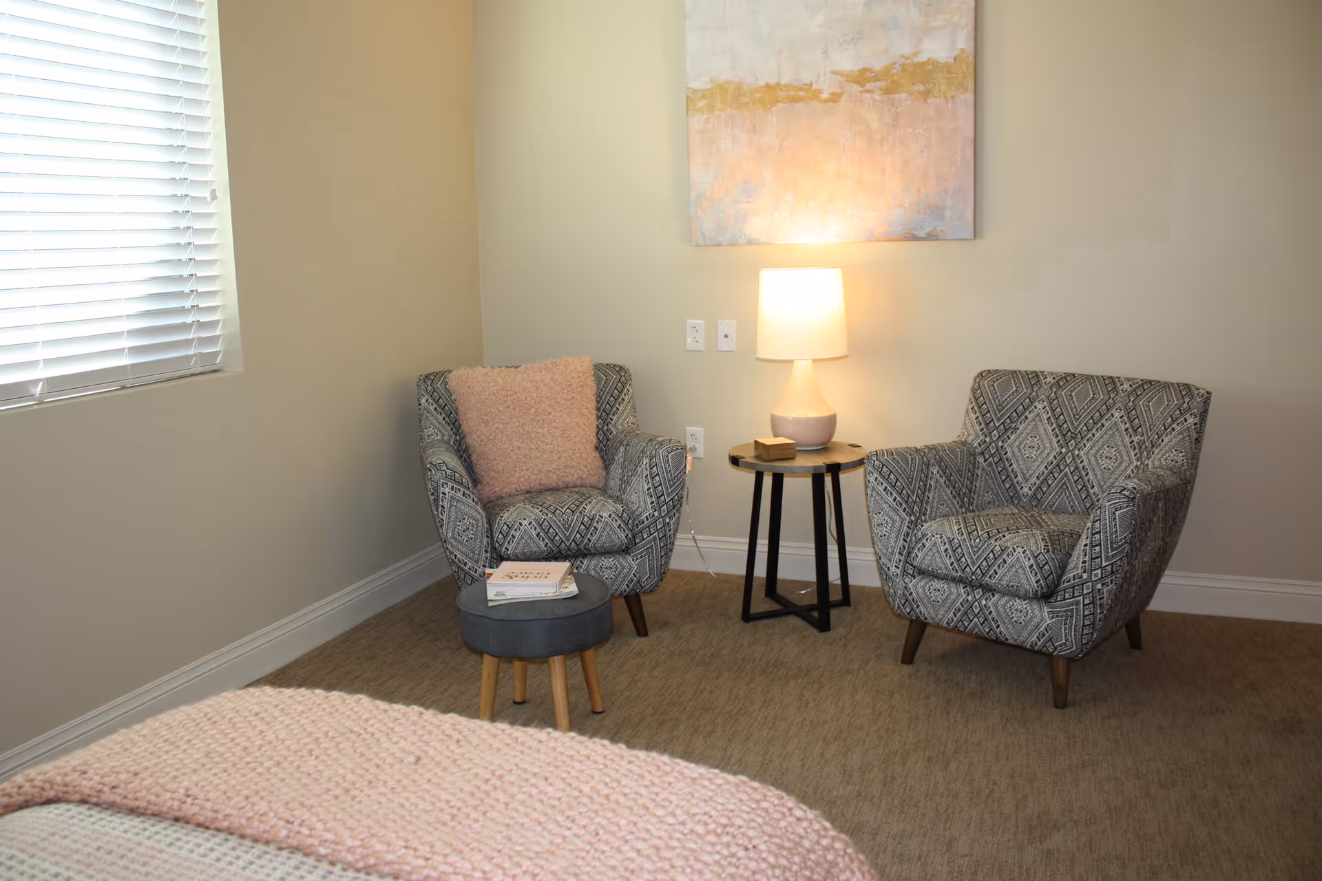 A cozy corner of a room featuring two patterned armchairs with wooden legs, a small round side table with a lamp, a small round ottoman with books on it, a window with blinds, and a piece of abstract artwork on the wall. The room has beige walls and carpeted flooring.