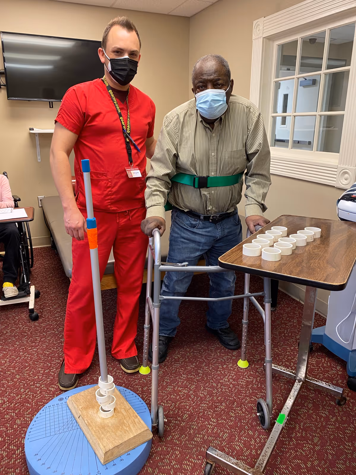 A healthcare worker in red scrubs and a black face mask stands next to an elderly man using a walker and wearing a green gait belt and a blue face mask. They are in a room with a red carpet, a TV mounted on the wall, and a table with several white rings on it, likely for a therapy or rehabilitation activity.