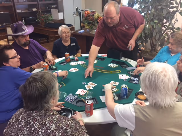 A group of elderly people sitting around a green poker table playing cards and using poker chips. A man in a maroon shirt is standing and reaching over the table, possibly dealing or collecting chips. The room has shelves, plants, and decorations in the background.