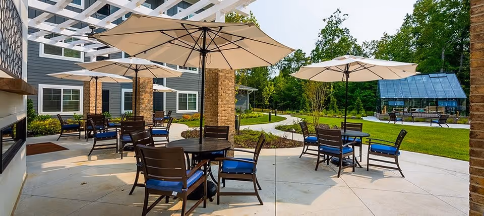 Outdoor patio with round tables, blue-cushioned chairs and large umbrellas next to a building and lawn.