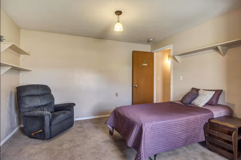 A simple bedroom with a single bed covered in a purple bedspread and two pillows. There is a dark gray recliner chair on the left side and a wooden nightstand next to the bed. The walls are beige with white shelves mounted on two sides. A wooden door is open, revealing a lit hallway.