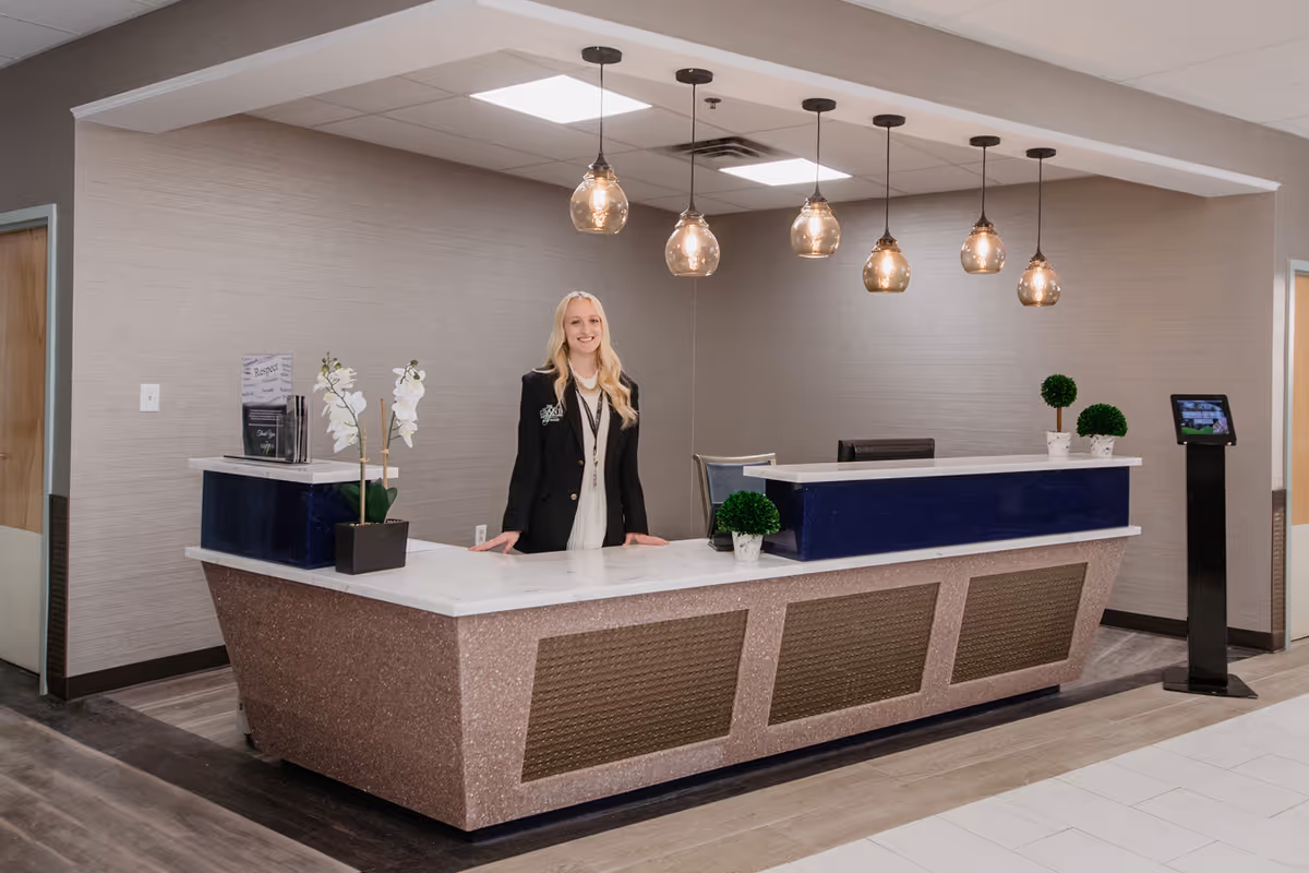 A woman standing behind a modern reception desk in a well-lit interior space. The desk has a marble countertop with decorative plants and hanging pendant lights above. The background features neutral-colored walls and a doorway on the left side.