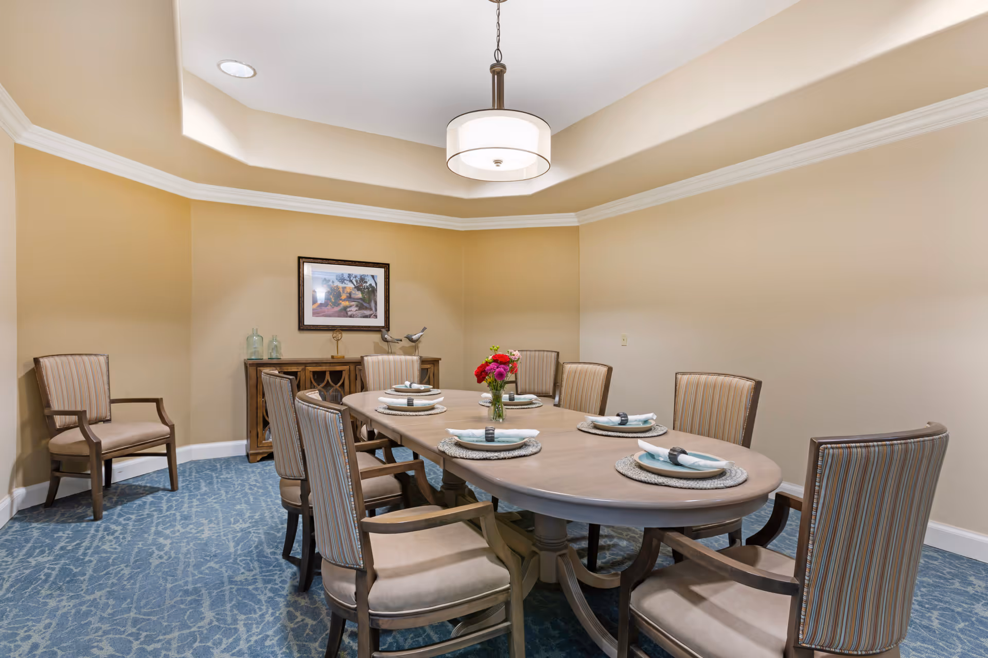 A dining room with a long oval table set for six people with plates, napkins, and a vase of flowers in the center. The room has beige walls, a blue patterned carpet, a ceiling light fixture, and a sideboard with decorative items and a framed picture above it.