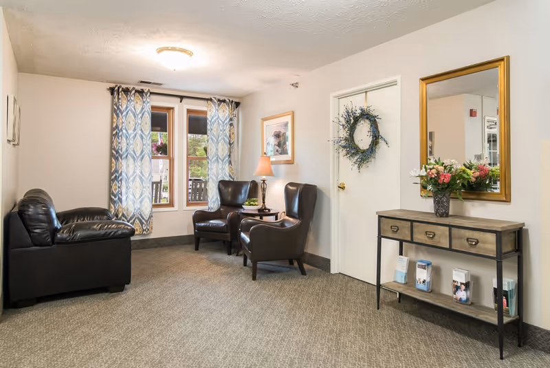 A cozy sitting area with two dark leather armchairs and a small table with a lamp between them, a black leather loveseat, a window with patterned blue and white curtains, a door decorated with a wreath, and a console table with a vase of flowers and brochures underneath a large mirror.