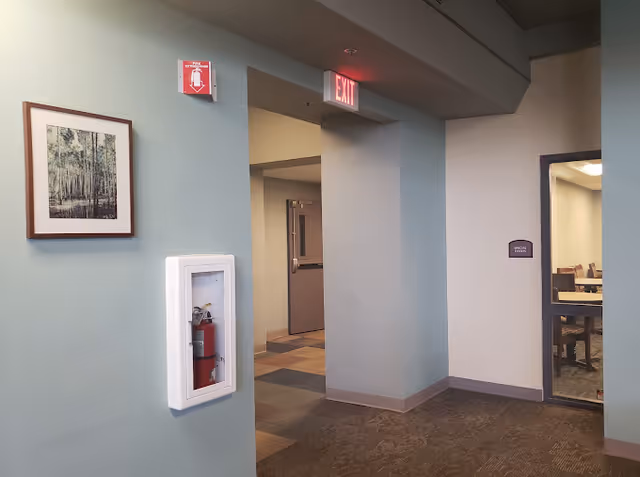 Interior hallway of a facility with light blue walls, a framed picture of trees on the left wall, a fire extinguisher in a white recessed cabinet, an exit sign above a doorway, and a room with tables and chairs visible through a glass window on the right.
