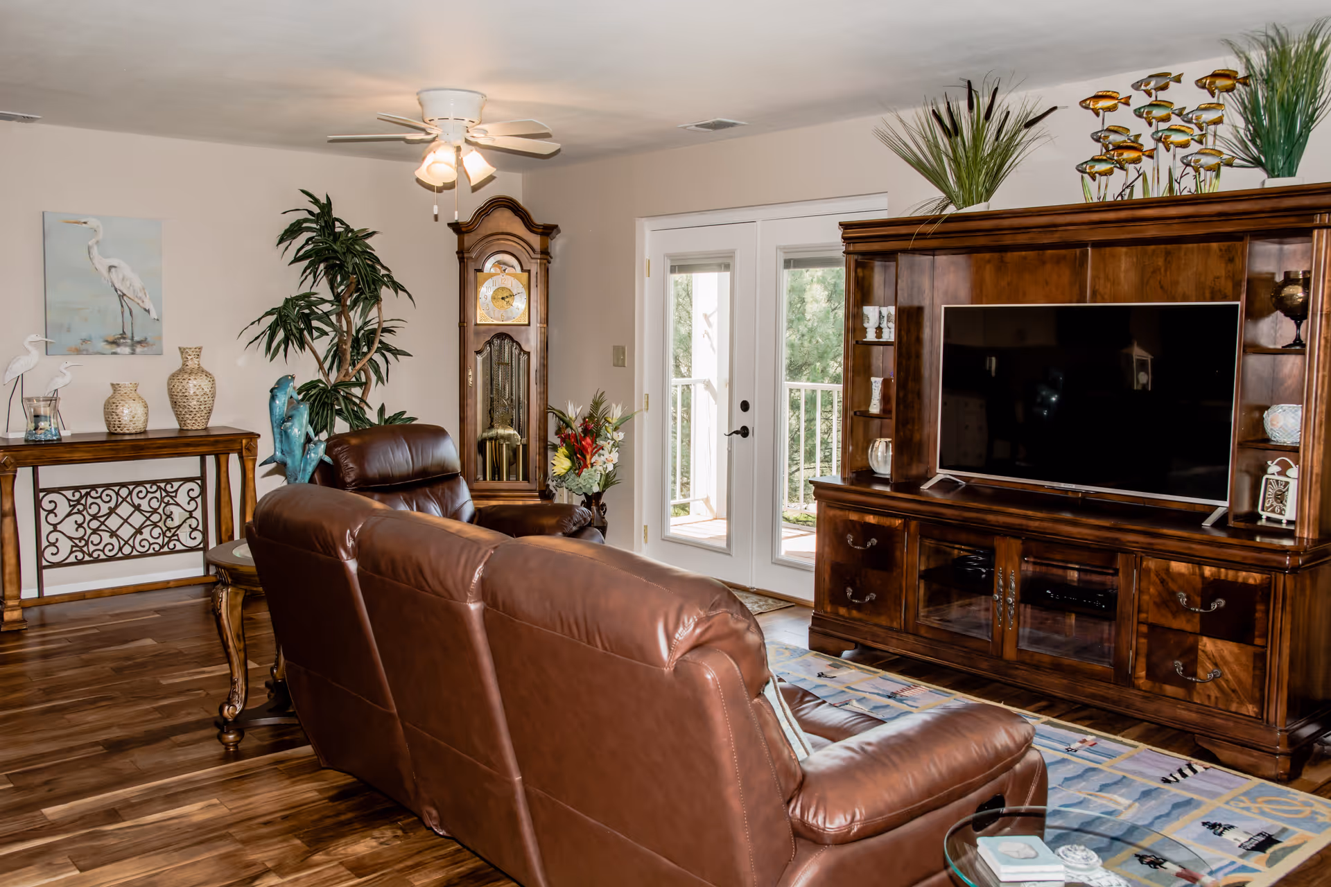 A cozy living room with a brown leather sofa facing a large wooden entertainment center with a flat-screen TV. The room features a wooden floor, a grandfather clock, a ceiling fan with lights, decorative plants, and a console table with vases and bird figurines. French doors lead to an outdoor balcony.