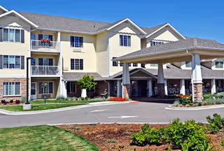 Exterior view of a multi-story retirement community building with beige siding and stone accents. The building features balconies, a covered entrance with pillars, and landscaped grounds with green grass and shrubs.
