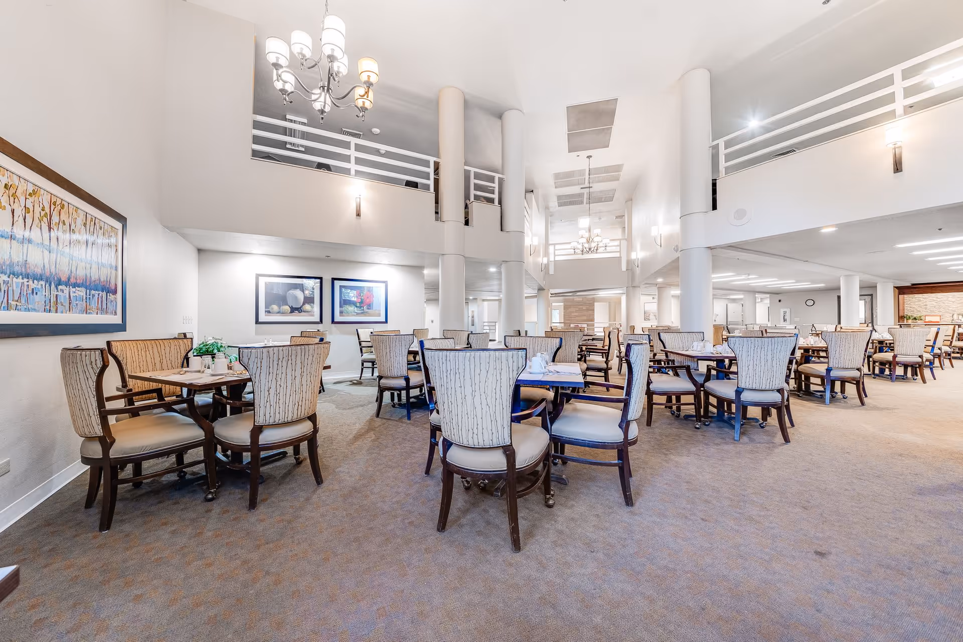 Spacious dining room with multiple tables and cushioned chairs arranged neatly. The room features high ceilings with chandeliers, white walls, and framed artwork. There is a balcony railing visible on the upper level and soft lighting throughout the area.