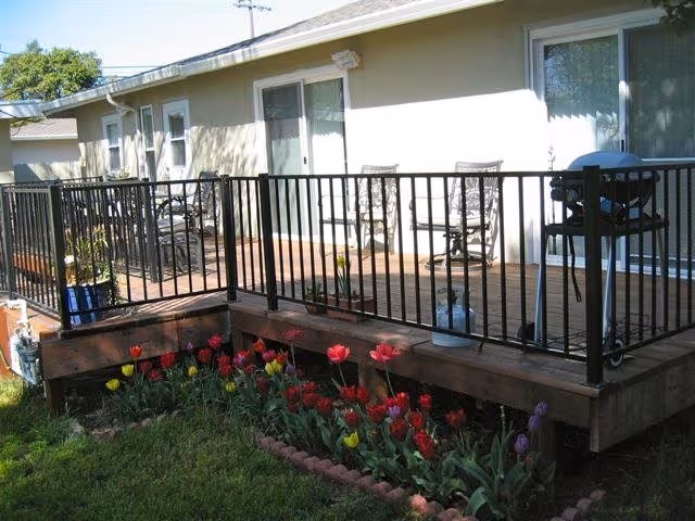 Exterior view of a single-story building showing a wooden deck with black metal railing, patio chairs and a grill, and a tulip flowerbed in front.
