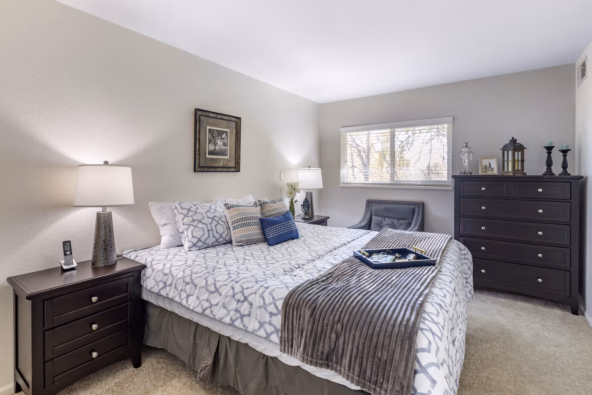 A neatly arranged bedroom with a large bed covered in patterned bedding and multiple pillows. There are two dark wooden nightstands on either side of the bed, each with a lamp. A window with blinds is on the right wall, letting in natural light. A dark wooden dresser with decorative items on top is placed against the right wall, and a cushioned chair is positioned near the window. The walls are painted light gray, and a framed picture hangs above the bed.