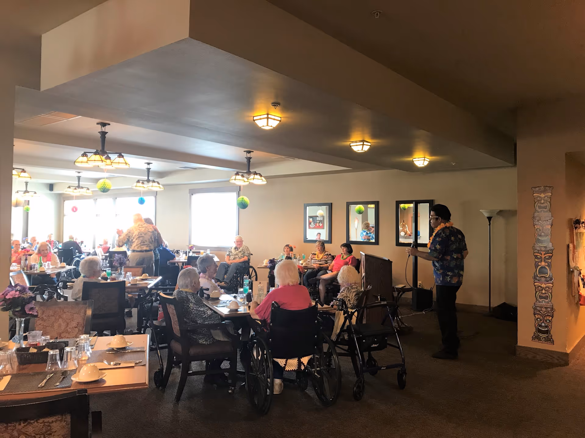 A dining room in an assisted living facility with elderly residents seated at tables, some in wheelchairs. A man stands near a podium holding a microphone, possibly addressing the group or performing. The room is warmly lit with ceiling lights and decorated with hanging paper ornaments and wall art.