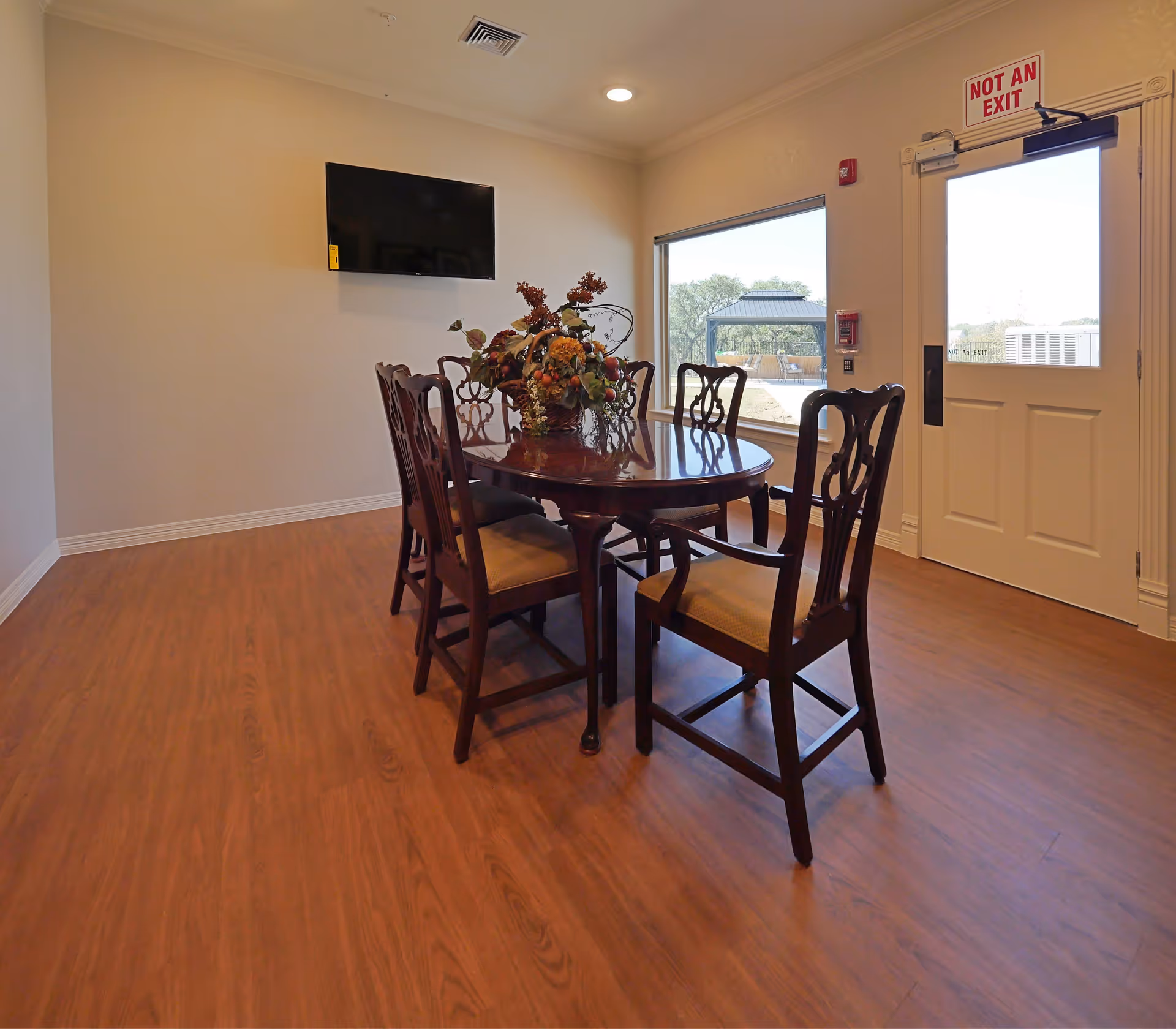 A small dining area with a wooden oval table and six matching chairs with cushioned seats. A floral centerpiece decorates the table. A flat-screen TV is mounted on the beige wall. There is a large window and a door with a 'NOT AN EXIT' sign above it. The floor is wood laminate.