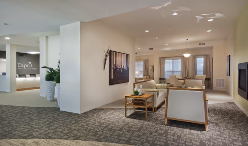A spacious and well-lit senior living facility common area with comfortable seating including sofas and armchairs arranged around a wooden coffee table. The room features neutral tones, carpeted floors, large windows with curtains, and a wall-mounted television. In the background, there is a reception desk with the sign 'Esprit Whispering Ridge' on the wall and decorative plants nearby.