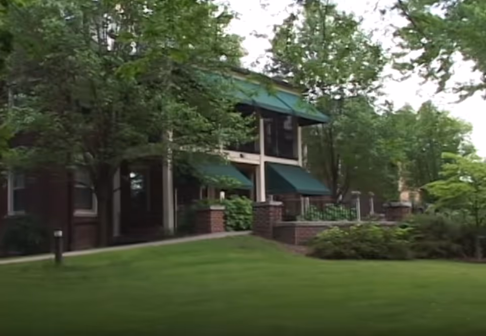 Exterior view of a building with green awnings over windows and a porch area, surrounded by trees and well-maintained green lawn.