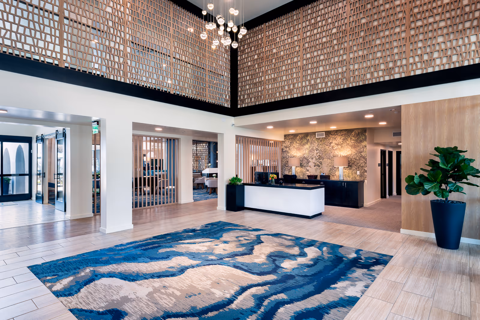 Spacious and modern lobby area with a large blue and beige patterned rug on the floor, a reception desk with two lamps and decorative wallpaper behind it, tall decorative wooden lattice panels near the ceiling, and a large potted plant on the right side.