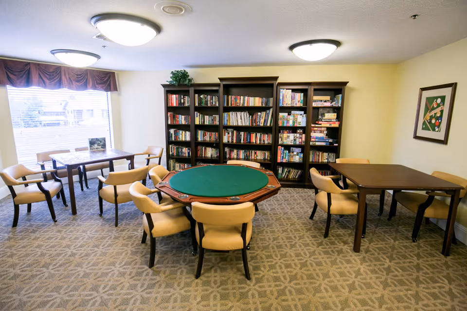 A bright communal activity room with a round green-felt card table, additional tables and chairs, and bookshelves along the back wall.