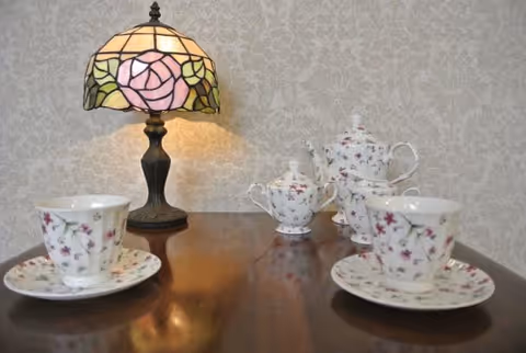 A wooden table set with floral china teacups, saucers and a teapot beside a stained-glass table lamp against patterned wallpaper.