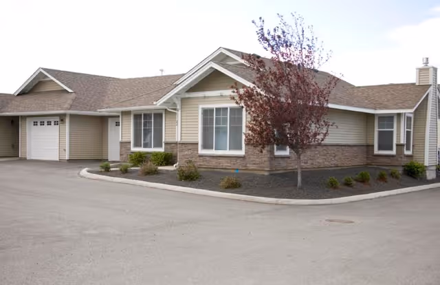 Exterior view of a single-story senior living facility building with beige siding, stone accents, a garage, and a small landscaped area with a tree and shrubs.
