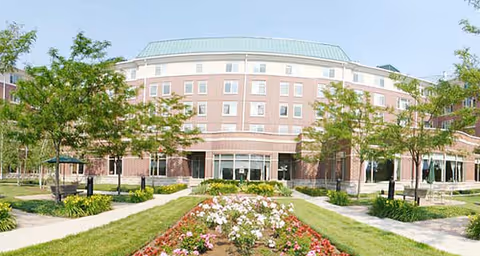 Front exterior view of a large, multi-story senior living facility with a curved facade, brick walls, and a green roof. The building is surrounded by landscaped gardens with trees, flower beds, and paved walkways.