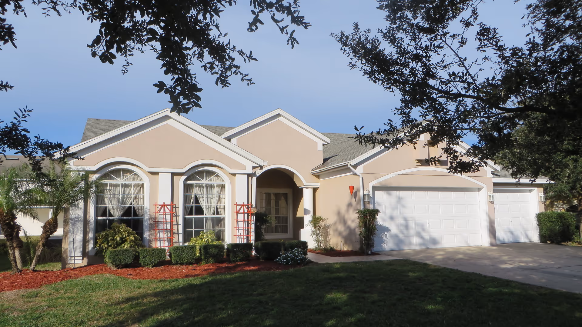 Single-story beige house with arched windows, an attached three-car garage, and landscaped front yard under a clear blue sky.