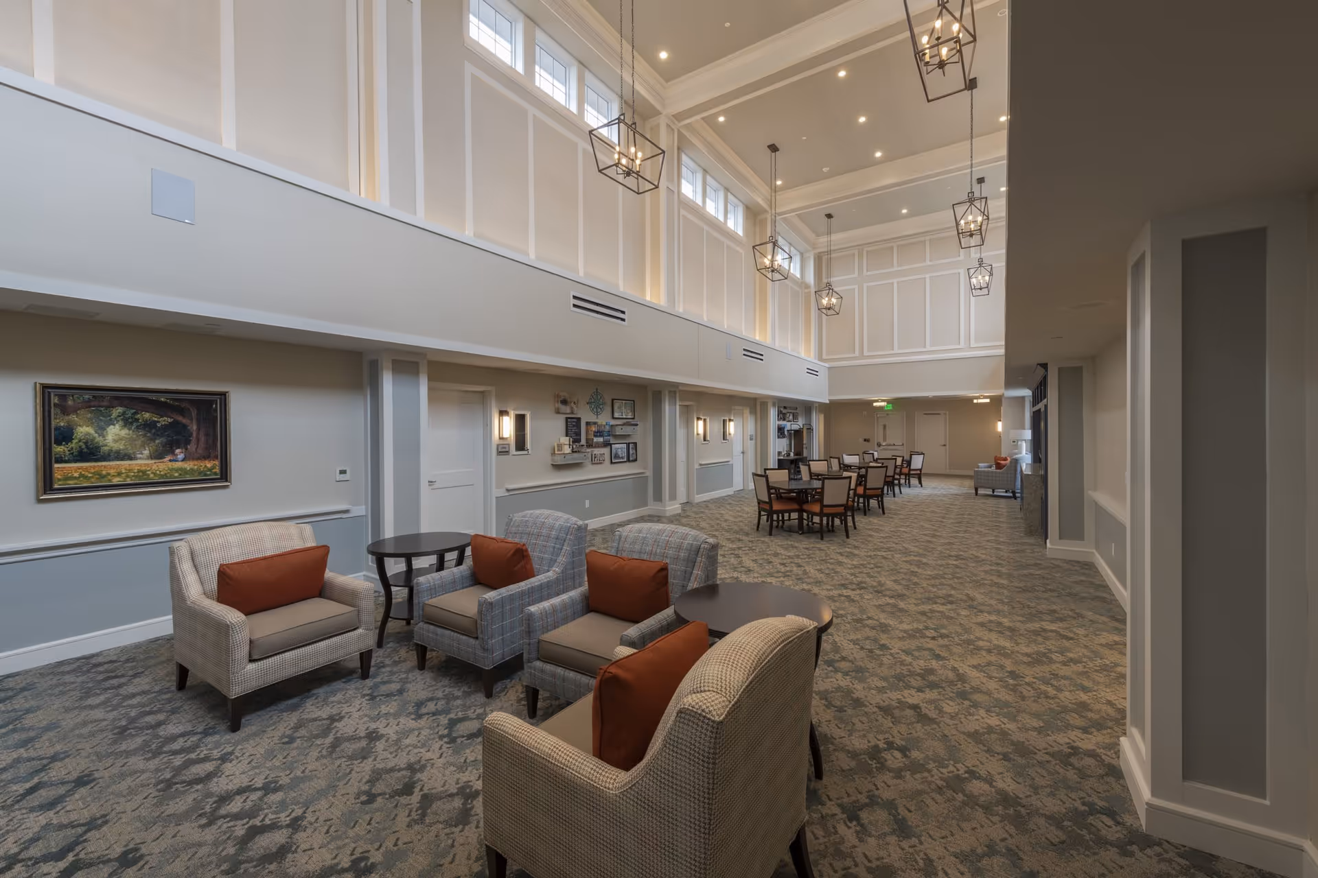 Spacious and well-lit common area with high ceilings and multiple hanging light fixtures. The room features several comfortable armchairs with orange cushions arranged around small round tables, and dining tables with chairs further back. The walls are decorated with framed artwork and decorative items, and there are large windows near the ceiling allowing natural light to enter.
