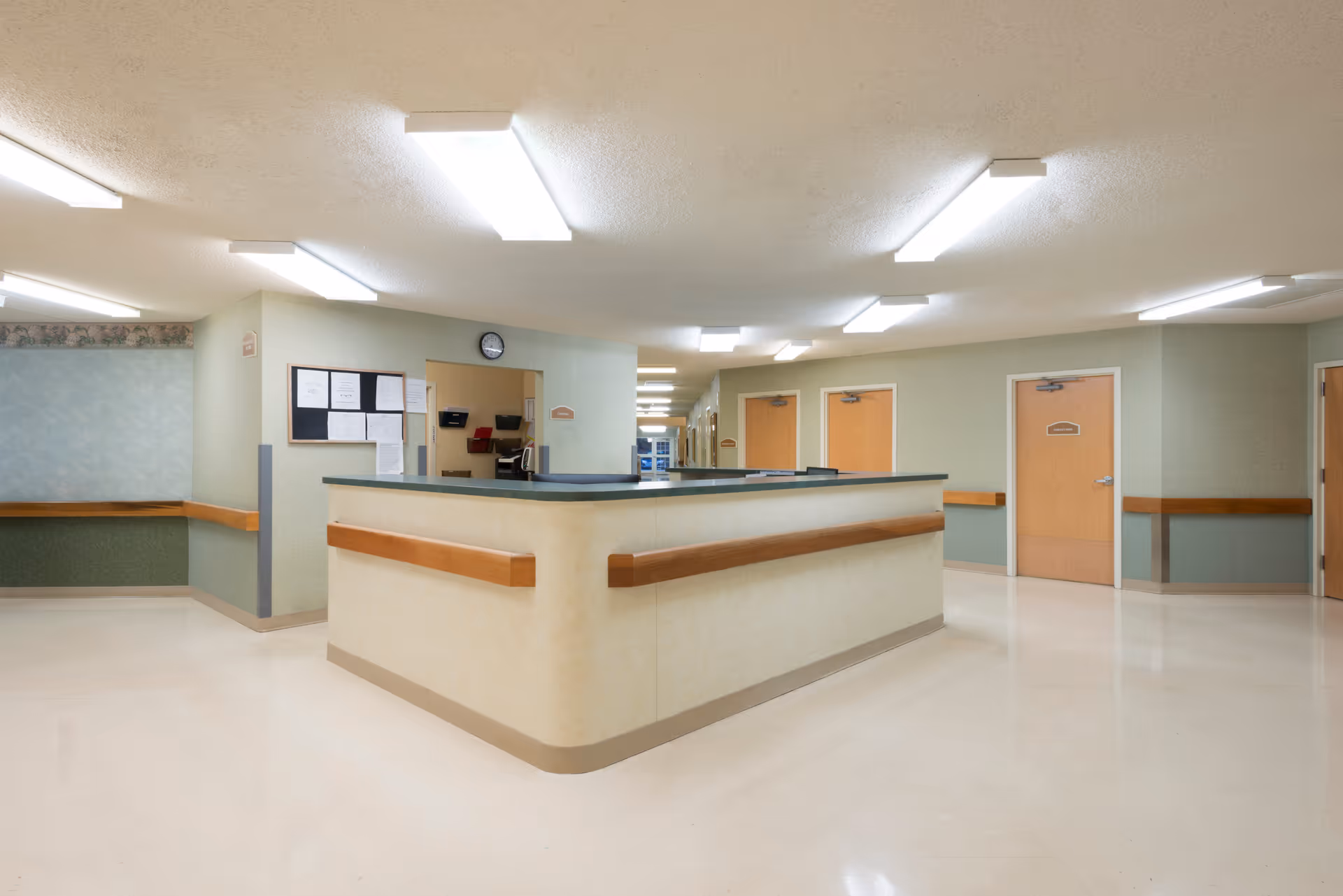 Well-lit interior corridor with a central nurses' station/reception desk, handrails, and several room doors.