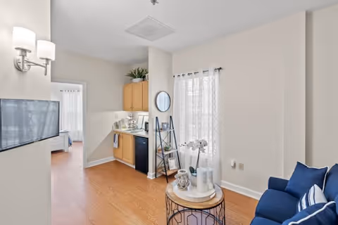 A bright and clean interior space featuring a small kitchenette with light wood cabinets, a black mini fridge, and a sink. To the right, there is a blue couch with striped pillows and a round coffee table decorated with candles and a white orchid. A window with sheer white curtains allows natural light to fill the room. In the background, a doorway leads to a bedroom with a bed visible. The floor is wooden, and the walls are painted white.