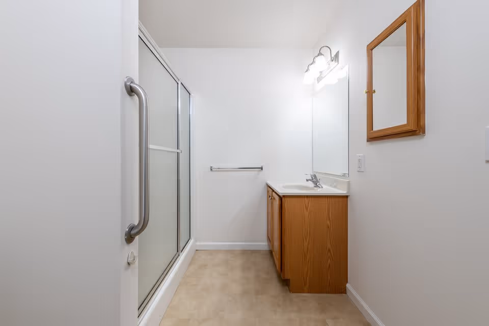 A clean bathroom featuring a shower with sliding frosted glass doors and a metal grab bar, a wooden vanity with a sink and faucet, a large mirror above the vanity, a wall-mounted wooden medicine cabinet, and a towel rack on the wall. The walls are white and the floor has a beige tile pattern.