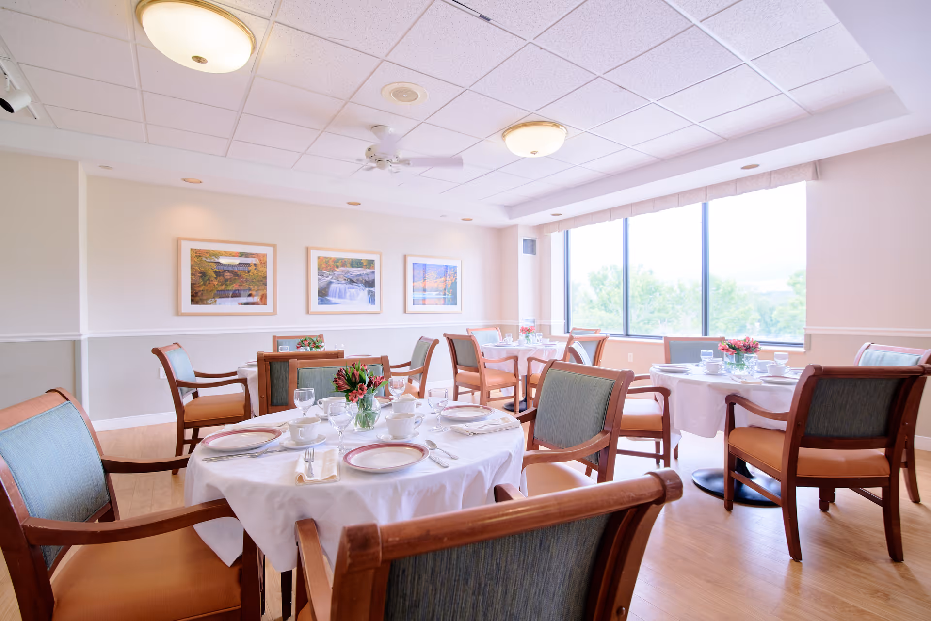 A bright dining room with several round tables covered in white tablecloths, each set with plates, cups, glasses, and silverware. Wooden chairs with green upholstery surround the tables. Large windows let in natural light and offer a view of greenery outside. Three framed landscape photographs hang on the far wall.