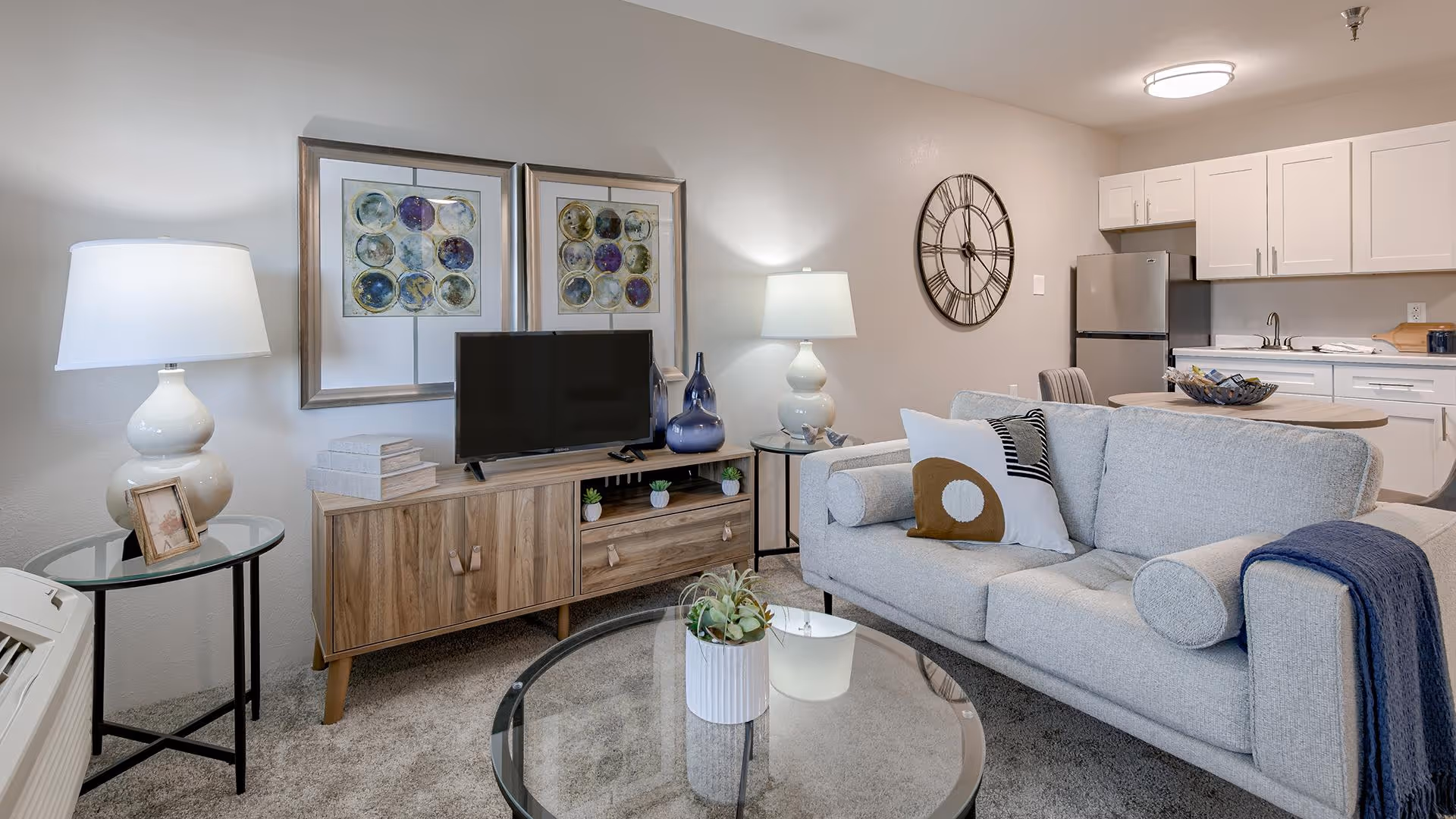 A modern living room and kitchen area in an independent living facility. The living room features a light gray sofa with decorative pillows and a blue throw blanket, a round glass coffee table with a small plant, and a wooden TV stand with a flat-screen TV and decorative items. Two framed abstract artworks hang on the wall above the TV stand. There is a side table with a white lamp and a small framed photo. The kitchen area in the background has white cabinets, a stainless steel refrigerator, a sink, and a round dining table with chairs. A large wall clock is mounted on the wall near the kitchen.