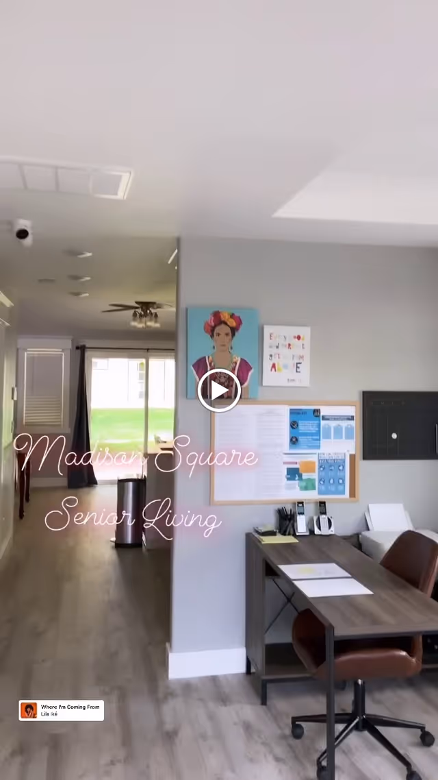 Interior view of a senior living facility office area with a desk, chair, bulletin board with notices, and artwork on the wall. A hallway with wood flooring leads to a bright room with large windows and a ceiling fan.