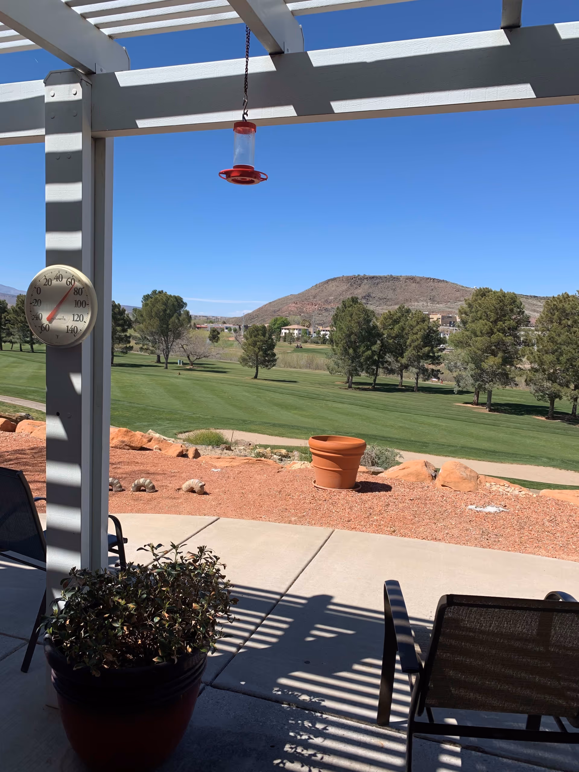 View from a shaded patio area with a pergola overhead, featuring a hanging bird feeder and a thermometer on a post. There are chairs and potted plants on the patio, with a landscaped area of red gravel and a large empty terracotta pot. Beyond the patio is a green lawn with trees and a hill in the background under a clear blue sky.