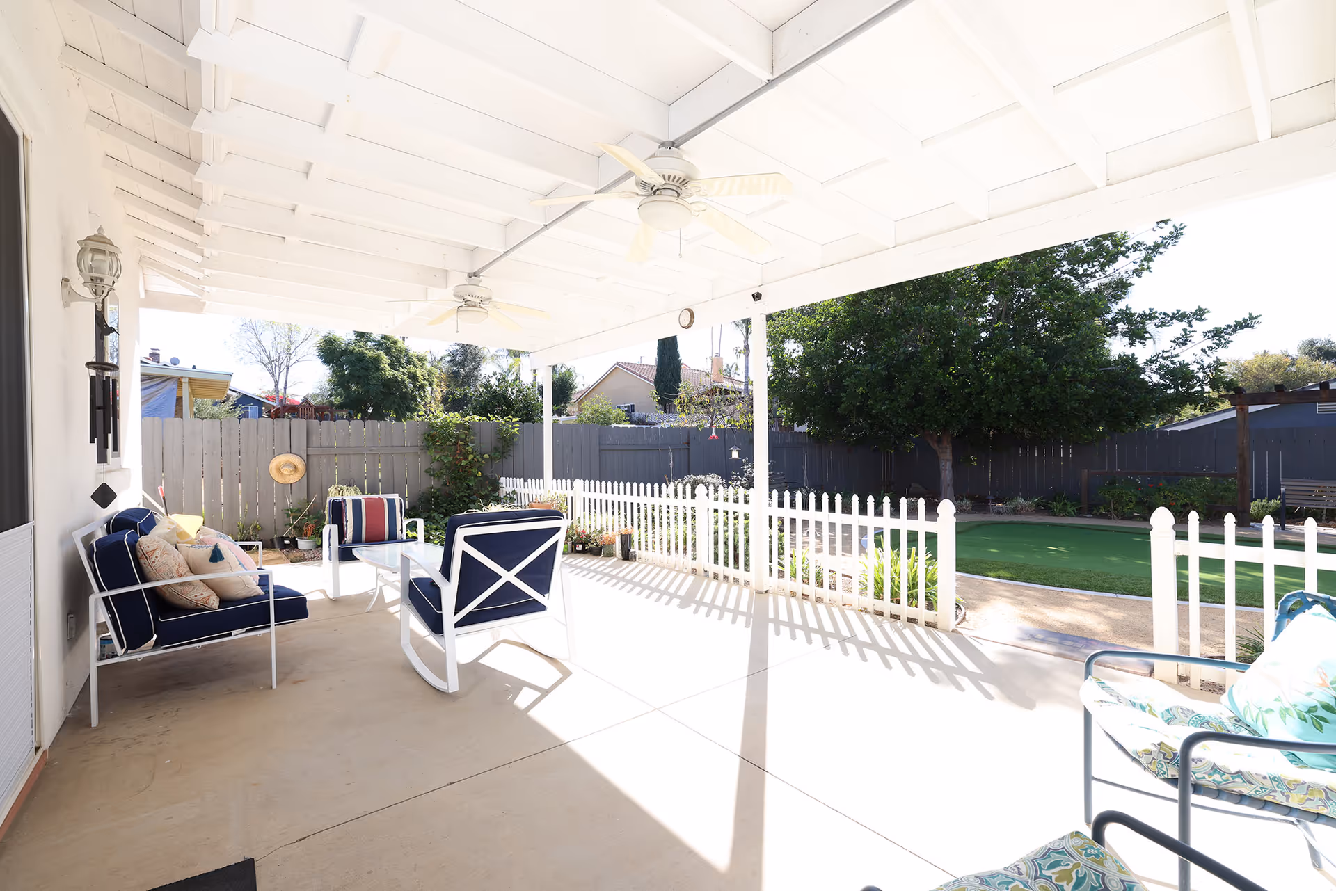 Covered outdoor patio area with white wooden ceiling and ceiling fans, furnished with cushioned chairs and a glass-top table. The patio overlooks a fenced backyard with green grass, trees, and a small putting green.