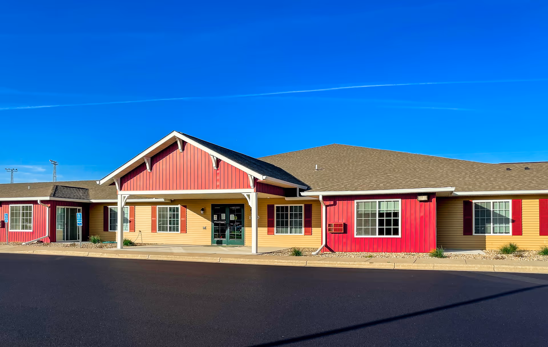 Exterior front view of a single-story building with a covered entrance. The building has red and beige siding with white trim and several windows. The sky is clear and blue above.