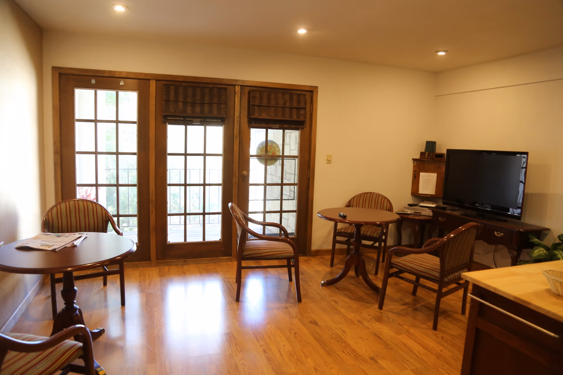 Sunlit common sitting area with wooden floors, French doors, round tables, striped chairs, and a TV on a cabinet.
