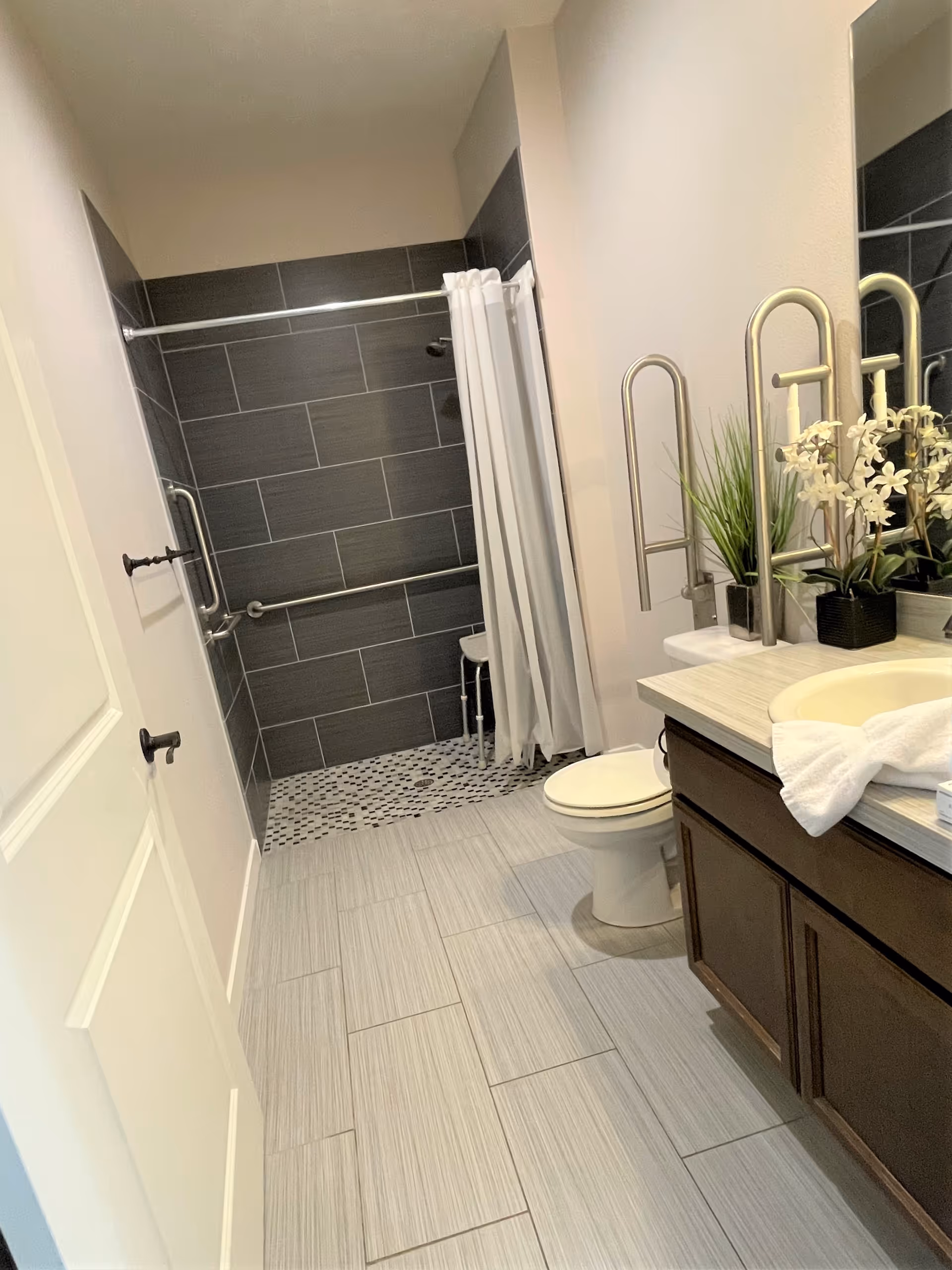 A modern bathroom with light gray floor tiles and dark gray tiled walk-in shower area with a white shower curtain. The bathroom features a white toilet, a wooden vanity with a white countertop, a sink, and decorative plants next to a large mirror. There are safety grab bars installed near the shower and sink.