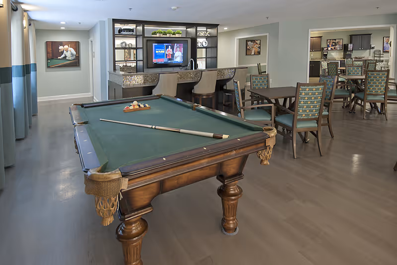 A spacious common area in a senior living facility featuring a pool table with a cue stick and balls arranged for a game. Behind the pool table is a bar counter with three high chairs and a mounted TV screen. To the right, there are several tables and chairs arranged for socializing or dining. The room has light-colored walls, wood flooring, and framed artwork on the walls.