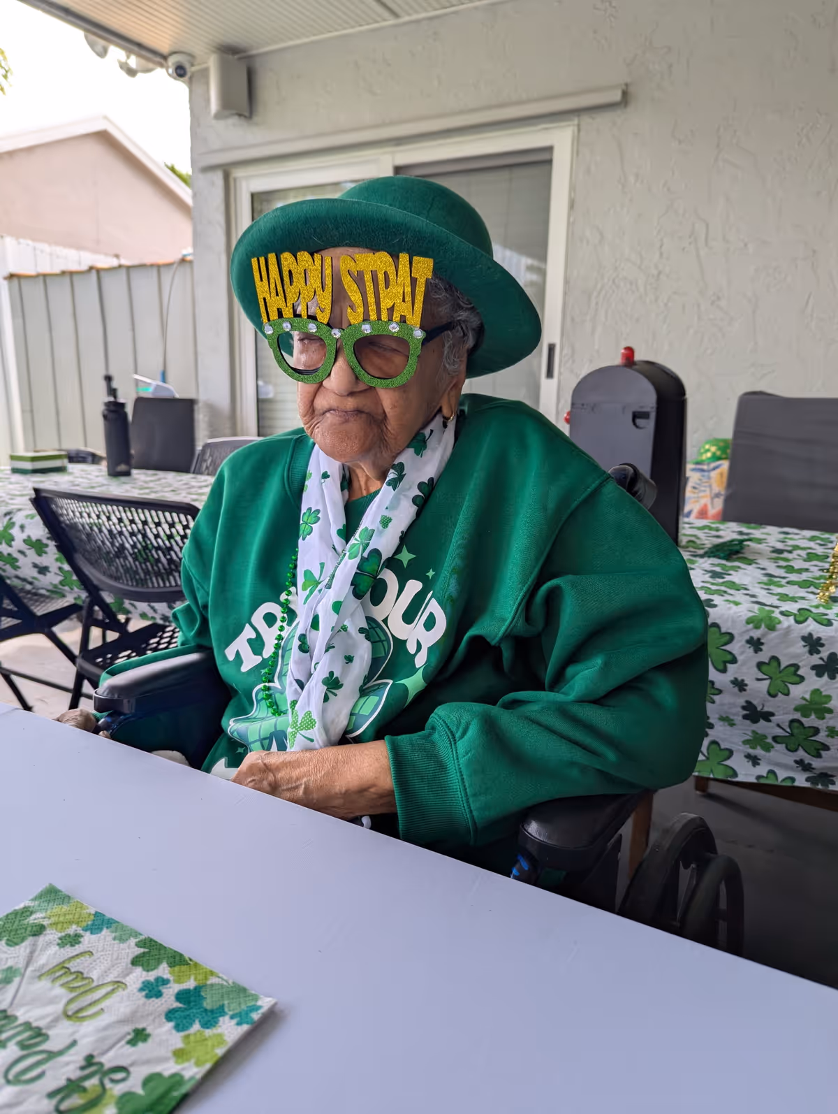 An elderly person wearing a green hat and large green glasses with the words 'HAPPY ST PAT' on them, dressed in a green sweatshirt and a white scarf with shamrock patterns, sitting in a wheelchair at a table decorated with shamrock-themed tablecloth and napkins, celebrating St. Patrick's Day on a covered patio.