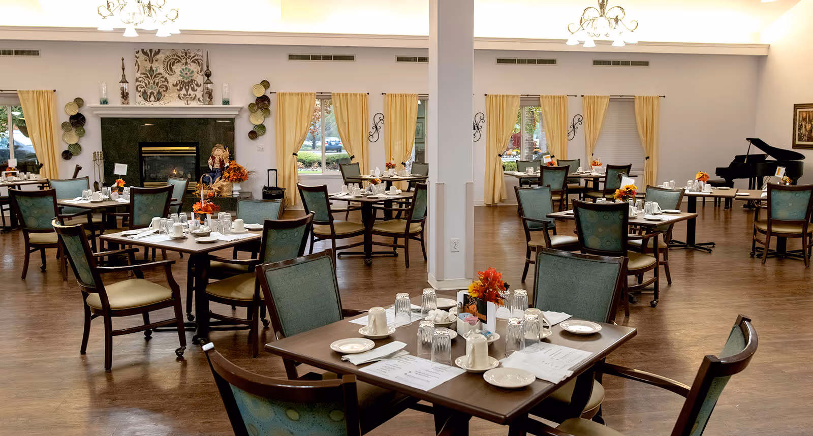 Spacious dining room with multiple set tables and chairs, a fireplace, piano, and seasonal floral centerpieces.