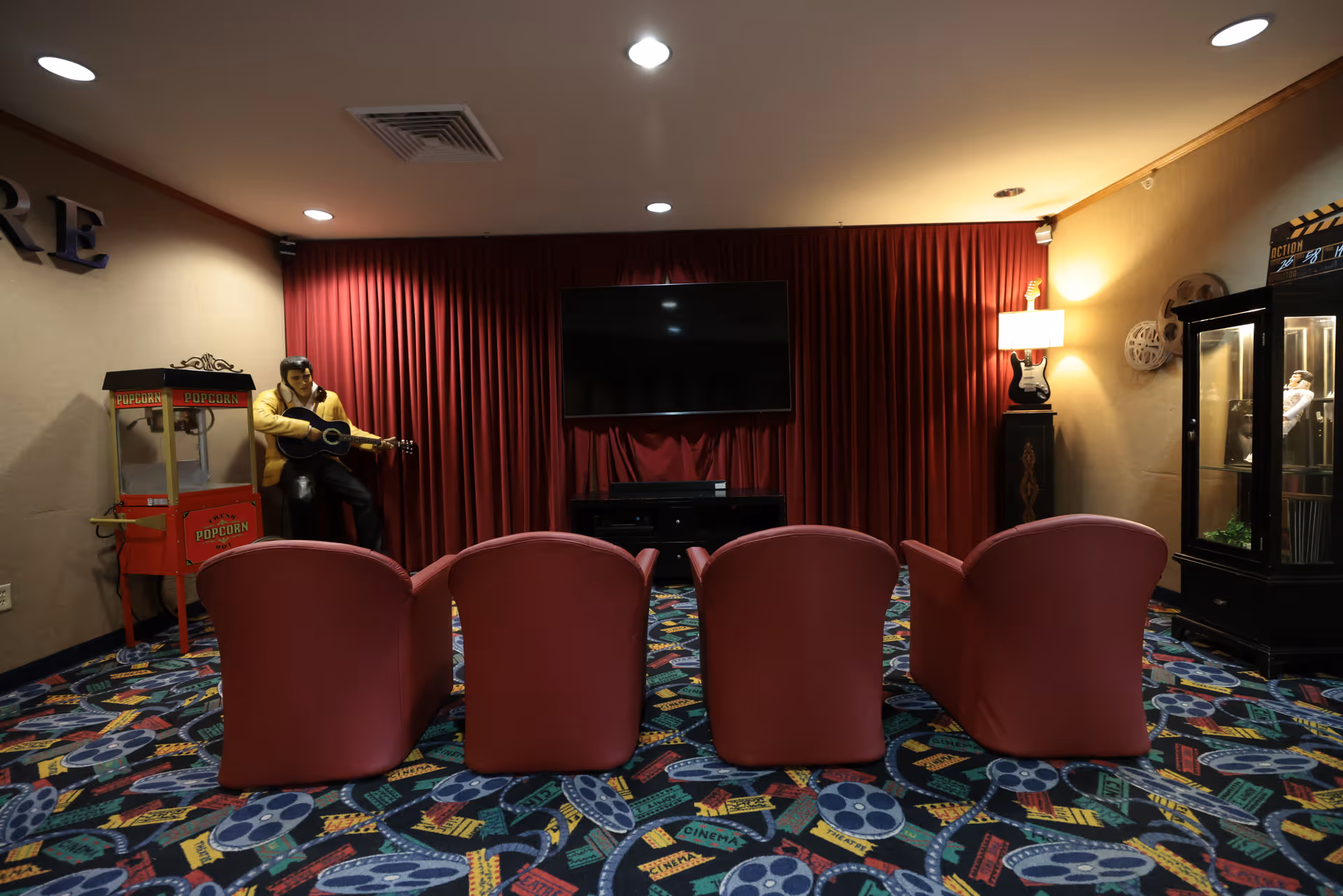 A small theater room with four red chairs facing a flat-screen TV mounted on a red curtain backdrop. To the left, there is a popcorn machine and a life-size statue of a man playing a guitar. On the right side, there is a display cabinet with memorabilia and a lamp shaped like a guitar. The carpet has a colorful cinema-themed pattern.